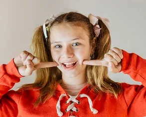 Smiling girl in an orange sweatshirt showcasing her teeth with fingers, representing the joy of orthodontic treatment and healthy smiles for children, relevant to Gellerman Orthodontics' focus on Invisalign First.
