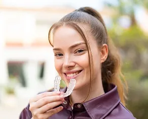 Teen girl smiling while holding clear Invisalign aligners, showcasing a comfortable and discreet teeth straightening option, relevant to Gellerman Orthodontics' Invisalign services for teens in Huntington, NY.